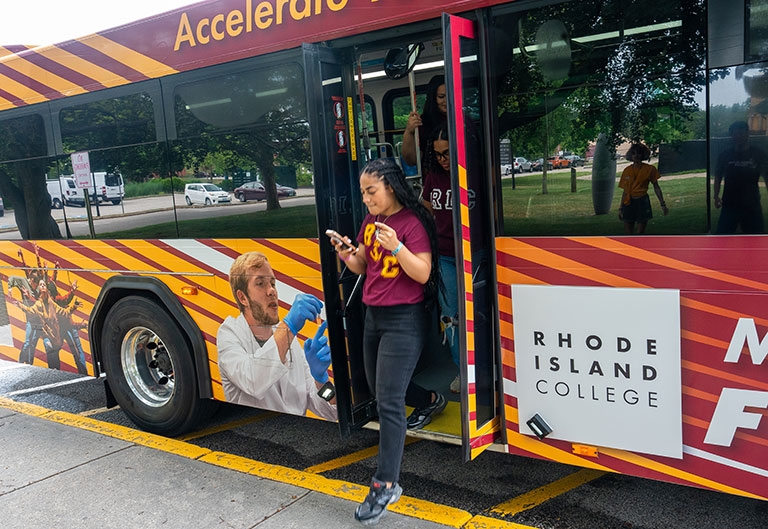 Student in RIC gear stepping off RIPTA, RIC-wrapped bus