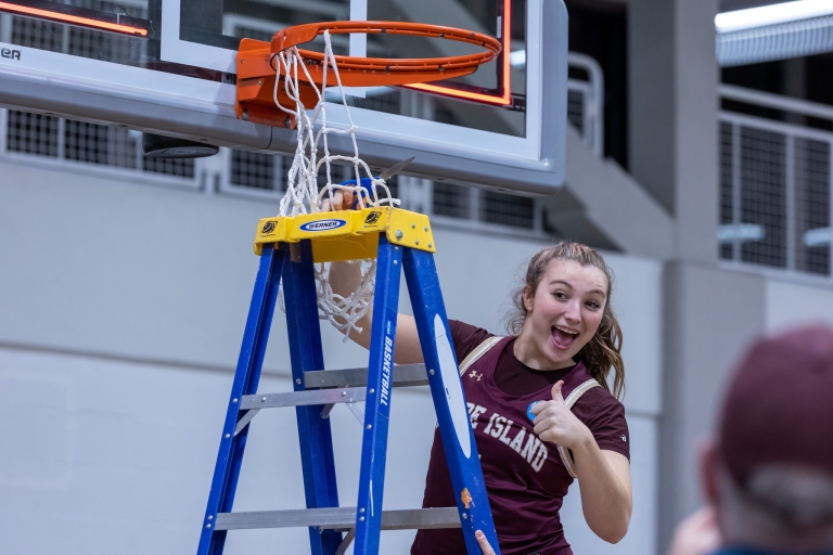 A RIC Women's Basketball player stands on a ladder to cut down the net from the basket after a victory.