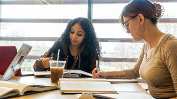 Female students at work at a table with books, notebooks, laptop, and coffee