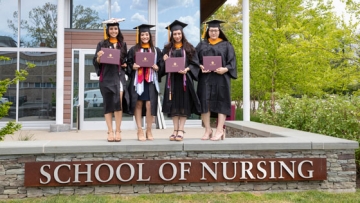 Nursing graduates in cap and gown with diplomas in front of the School of Nursing