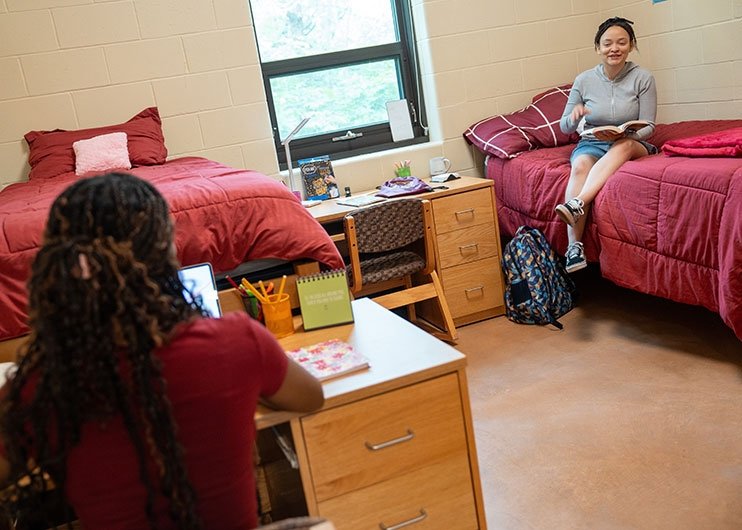 Students in their dorm room at a desk and sitting on bed with a book