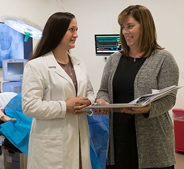 Person in lab coat speaking with another person, holding clipboard, in medical room