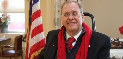 Congressman James Langevin seated in his office wearing a red scarf in front of an American flag
