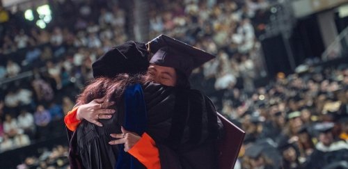 Graduate in cap and gown at ceremony in heartfelt embrace with robed faculty member