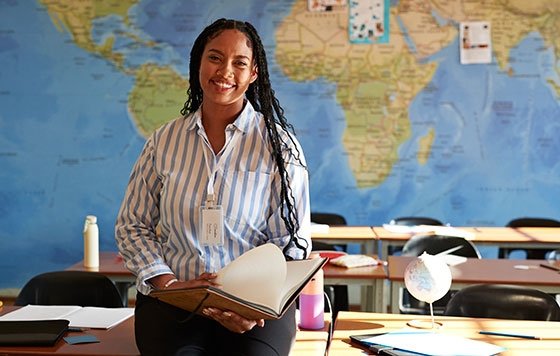 Black teacher standing at front of classroom with map of world, Africa prominent, behind her