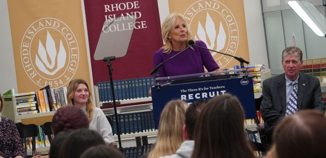 First Lady of the United States Dr. Jill Biden speaks in the Adams Library at Rhode Island College. She stands at a podium in front of three RIC banners. Governor Dan McKee is seated to her left. RIC student Grace Wells-Dannenfelser is seated at her right.